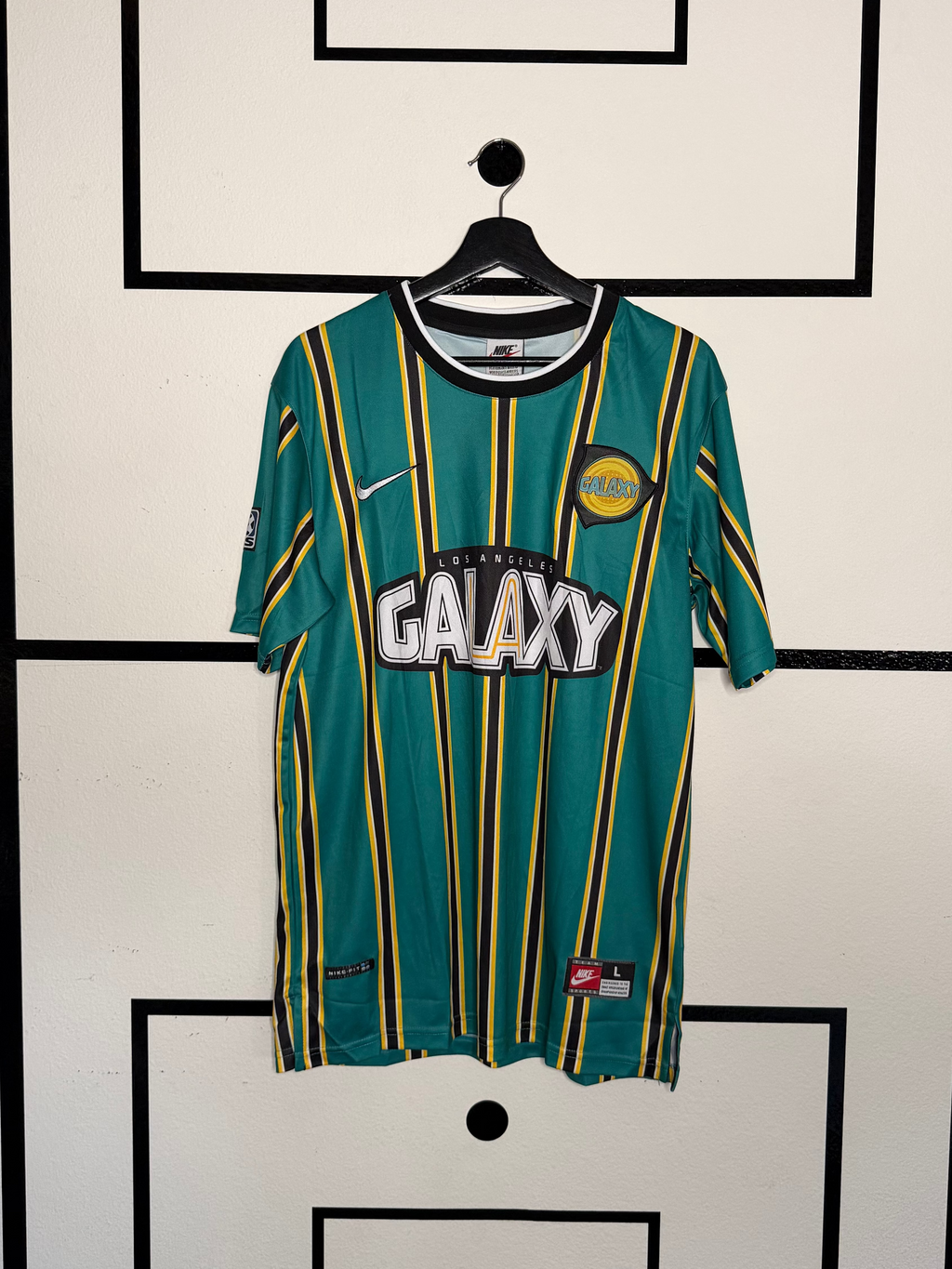 Green and yellow sports jersey with 'Galaxy' branding on a hanger against a white wall with black geometric patterns.