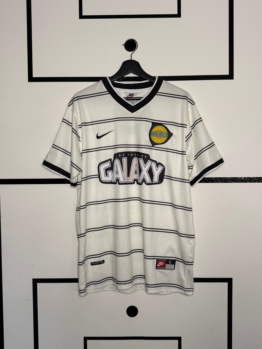 White sports jersey with black stripes and branding on a hanger against a wall with soccer field design
